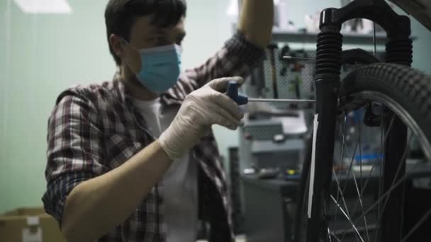 Механический ремонт велосипеда в мастерской с медицинским ковидом маски 19. Человек чинит велосипед на карантинной коронавирусной пандемии в защитной маске для лица. Человек исследует и исправит цикл. Концепция магазина — стоковое видео