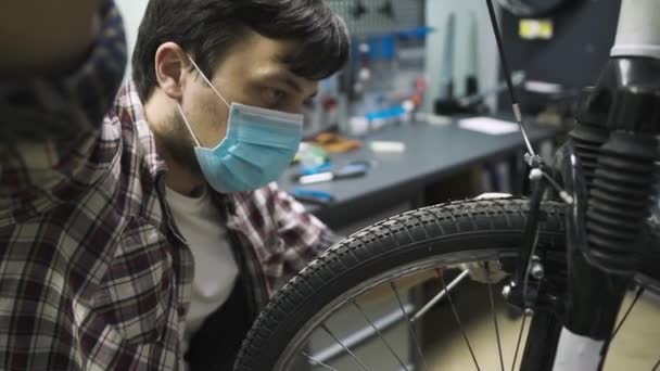 Tema mantenimiento de servicio y reparación de bicicletas en tienda de bicicletas durante la epidemia de coronavirus. Mecánica reparaciones bicicleta de montaña con máscara protectora, nuevas reglas covid 19. Asunto profesión de trabajador — Vídeo de stock