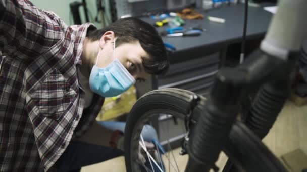 Mecánica de reparación de bicicletas en taller con máscara médica covid 19. Hombre bicicleta de reparación en cuarentena pandemia coronavirus en máscara facial protectora. El hombre examina y arregla el ciclo. Concepto tienda deporte — Vídeo de stock