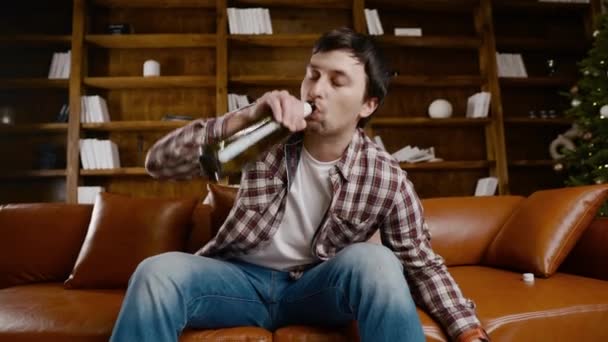 Депрессивный молодой человек пьет алкоголь дома. Безработный мужчина, пьян и продолжает пить вино прямо бутылку на диване возле елки. Добавление, одиночество, нервный срыв новогодних каникул — стоковое видео