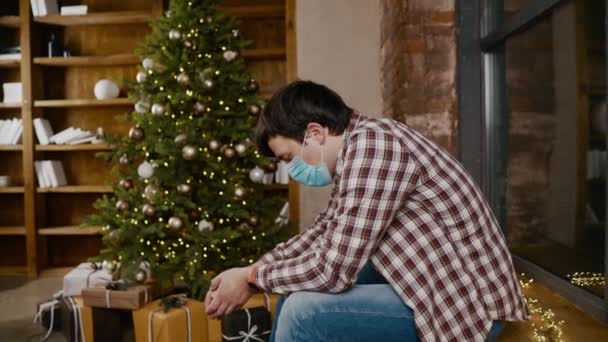 Depression during coronavirus epidemic. Lonely upset with bad mood young man in protective mask on Christmas evening near Christmas tree without friends and family. Lockdown Covid 19 and New Years — Stock Video