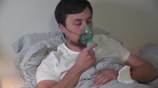 L'uomo respira attraverso la maschera per inalazione utilizzando il dispositivo medico di casa trattare le malattie del tratto respiratorio superiore e inferiore, asma e allergie a casa mentre si trova a letto durante la pandemia di coronavirus covid 19 — Video Stock