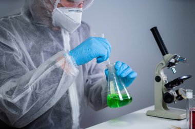Bilim adamı yeşil sıvıyı kabın içinde sallıyor. Laboratuvarda test ya da araştırma yapan genç bir laboratuvar asistanı. Teknisyen kimya laboratuarı deneyini test ediyor ve mikroskop ile birlikte cam tüpü sallıyor..