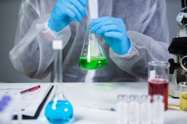 Erlenmeyer şişesinde kimya laboratuvarında yeşil sıvıyı karıştırıp sallayan koruyucu giysi teknisyeni. Laboratuvar teknisyeni kimyasal reaksiyonlar üzerinde çalışıyor ve kanser ilacı arıyor.