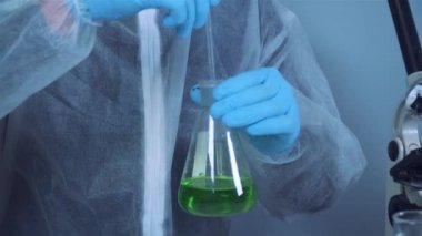 Bilim adamı yeşil sıvıyı kabın içinde sallıyor. Laboratuvarda test ya da araştırma yapan genç bir laboratuvar asistanı. Teknisyen kimya laboratuarı deneyini test ediyor ve mikroskop ile birlikte cam tüpü sallıyor.