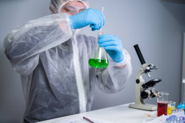 Bilim adamı yeşil sıvıyı kabın içinde sallıyor. Laboratuvarda test ya da araştırma yapan genç bir laboratuvar asistanı. Teknisyen kimya laboratuarı deneyini test ediyor ve mikroskop ile birlikte cam tüpü sallıyor..