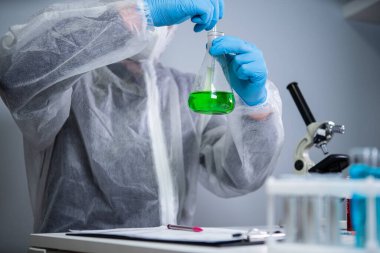 Erlenmeyer şişesinde kimya laboratuvarında yeşil sıvıyı karıştırıp sallayan koruyucu giysi teknisyeni. Laboratuvar teknisyeni kimyasal reaksiyonlar üzerinde çalışıyor ve kanser ilacı arıyor.
