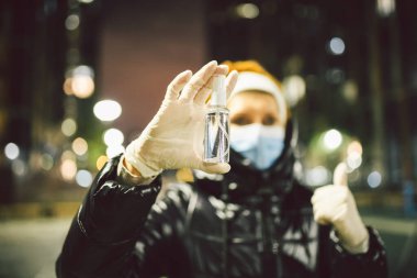 Koronavirüs ve kovid 19 'u koruyun. Kışın akşam vakti sokaklarda dolaşan olgun bir kadının elinde enfeksiyona karşı antiseptik. Maskeli yaşlı kadın ve ballı eldiven. Hastalık kapmaktan korkuyor..