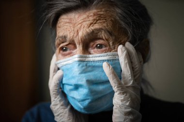 El ele tutuşan yaşlı bir kadının portresi koruyucu maske takan üzgün bir duygu, virüse karşı eldivenler ve evdeki enfeksiyona karşı. Yaşlı beyaz saçlı kadın karantinası evde kalır, yalnızlık ve korku.