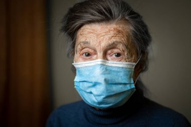 Yüzünde tıbbi maske olan yaşlı bir kadının akıllı görünüşü sizi mesafeli olmaya ve korunma ekipmanlarını kullanmaya teşvik eder. Kameraya bakan yaşlının portresi, yaşlı bakımı.
