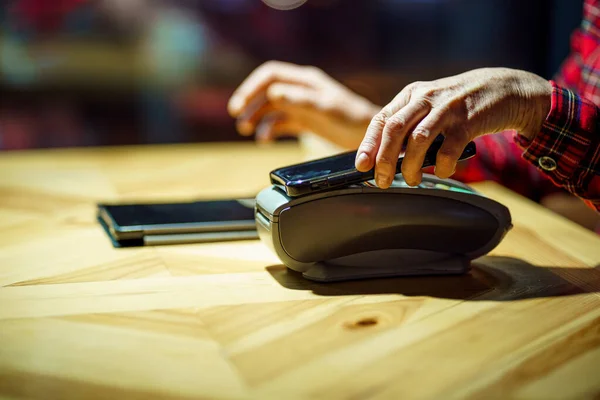 Female mature customer holding phone near nfc terminal make contactless mobile payment on coffeeshop counter, senior woman client pay in cafe with cellphone via pos machine. POS terminal.