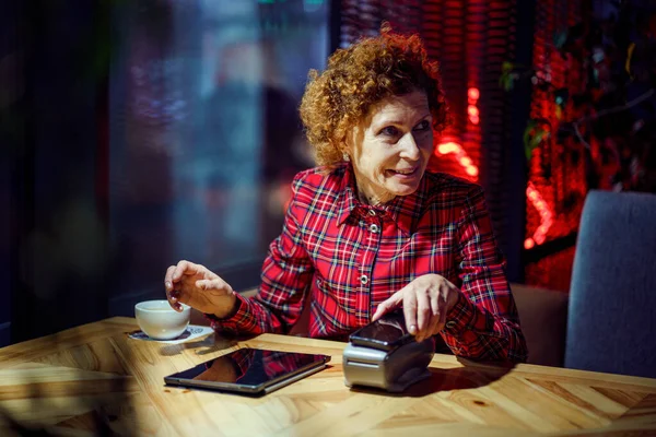 Female mature customer holding phone near nfc terminal make contactless mobile payment on coffeeshop counter, senior woman client pay in cafe with cellphone via pos machine. POS terminal.