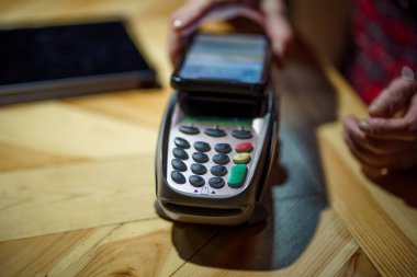 Olgun bir kadın, akşam kahve dükkanında bilgisayar terminali işlemleri ve akıllı telefon ödemeleri yapıyor. Üst düzey müşteri, kafenin parasını ödemek için akıllı telefonu kullanıyor. Kadın kafede ödeme yapıyor..