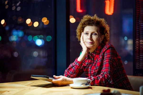 成熟的高加索女人喝咖啡 看电子片 晚上坐在市区咖啡馆里 老年女性在咖啡店使用技术 坐在有城市视野的橱窗旁 — 图库照片