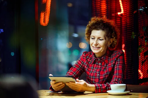 成熟した白人女性はコーヒーを飲み 電子タブレットを見て インターネットを使用します 夜はシティカフェの中に座って 高齢者の女性は街の景色と窓のそばに座ってコーヒーショップで技術を使用します — ストック写真