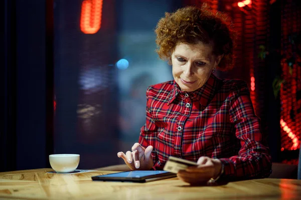 成熟的女人在咖啡店用平板电脑上网购物 老年妇女用信用卡支付网上付款 现代老年人在网上购物时使用塑料卡片 网上银行 — 图库照片