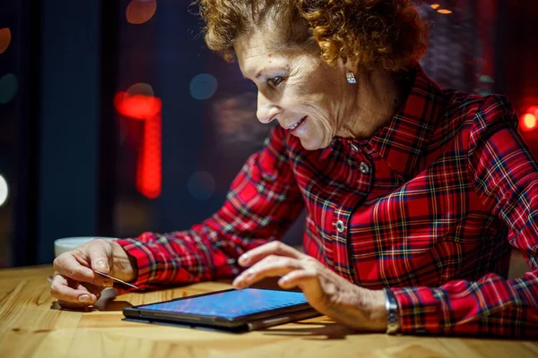 老年妇女在圣诞节网上购物 晚上坐在可以俯瞰城市的橱窗旁边的咖啡店里 成熟女人在网上用她的信用卡老年妇女在网上购物 — 图库照片