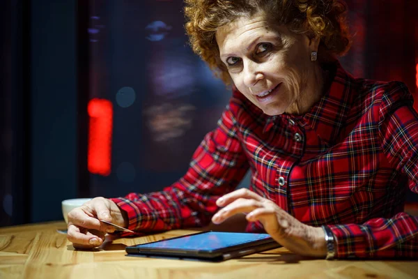 成熟的女人在咖啡店用平板电脑上网购物 老年妇女用信用卡支付网上付款 现代老年人在网上购物时使用塑料卡片 网上银行 — 图库照片