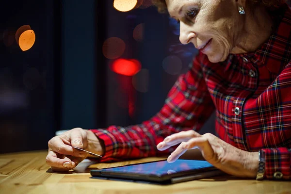 老年妇女在圣诞节网上购物 晚上坐在可以俯瞰城市的橱窗旁边的咖啡店里 成熟女人在网上用她的信用卡老年妇女在网上购物 — 图库照片