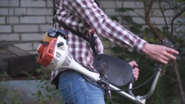 Elektrikli çim biçme makinesi ve çim biçme makinesi olan bir adam. Bahçıvan bahçe buduyor. Banliyö, köy. Beyaz tenli erkek bahçıvan avluda benzin makası ile dikiliyor. Tarlada yabani otları kesen fırça kesiciler