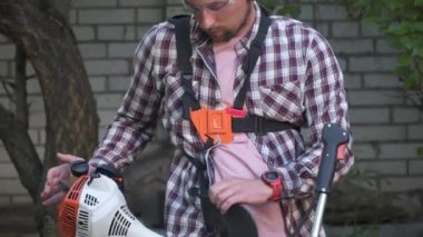 Genç Kafkasyalı çiftçi otları, uzun otları, çalıları ve çiftlik evinin arka bahçesindeki yabani otları budamak için benzinle çalışan elektrikli tırpan kullanıyor. Adam köyde çim biçme makinesiyle çalışıyor. Bahçe konsepti