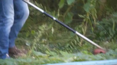 Bahçıvan kırsal evin bahçesinde çim biçme makinesiyle çalışıyor. Çiftlikte elektrikli tırpanla yabani otları kesen erkek çiftçi. Genç erkek işçi çim biçme makinesi kullanarak çim biçiyor. Benzin fırçası kesici.