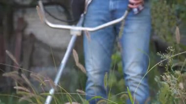 Elektrikli çim biçme makinesi ve çim biçme makinesi olan bir adam. Bahçıvan bahçe buduyor. Banliyö, köy. Beyaz tenli erkek bahçıvan avluda benzin makası ile dikiliyor. Tarlada yabani otları kesen fırça kesiciler