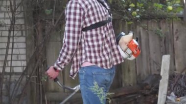 Bahçıvan kırsal evin bahçesinde çim biçme makinesiyle çalışıyor. Çiftlikte elektrikli tırpanla yabani otları kesen erkek çiftçi. Genç erkek işçi çim biçme makinesi kullanarak çim biçiyor. Benzin fırçası kesici.