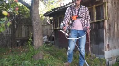 Elektrikli çim biçme makinesi ve çim biçme makinesi olan bir adam. Bahçıvan bahçe buduyor. Banliyö, köy. Beyaz tenli erkek bahçıvan avluda benzin makası ile dikiliyor. Tarlada yabani otları kesen fırça kesiciler