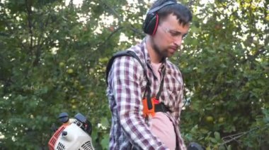 Genç Kafkasyalı çiftçi otları, uzun otları, çalıları ve çiftlik evinin arka bahçesindeki yabani otları budamak için benzinle çalışan elektrikli tırpan kullanıyor. Adam köyde çim biçme makinesiyle çalışıyor. Bahçe konsepti