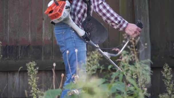 Elektrikli çim biçme makinesi ve çim biçme makinesi olan bir adam. Bahçıvan bahçe buduyor. Banliyö, köy. Beyaz tenli erkek bahçıvan avluda benzin makası ile dikiliyor. Tarlada yabani otları kesen fırça kesiciler — Stok video