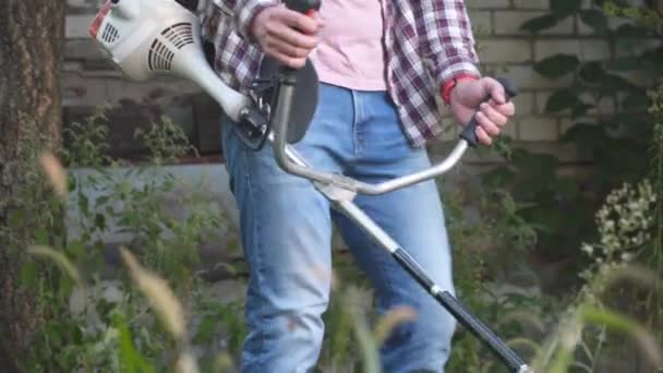若い白人農家は、農家の裏庭から雑草、背の高い草、茂みや雑草をトリミングするためにガソリン動力の電気鎌を使用しています。男は村の芝刈り機で働く。園芸の概念 — ストック動画