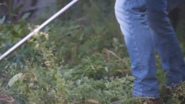 若い白人農家は、農家の裏庭から雑草、背の高い草、茂みや雑草をトリミングするためにガソリン動力の電気鎌を使用しています。男は村の芝刈り機で働く。園芸の概念 — ストック動画