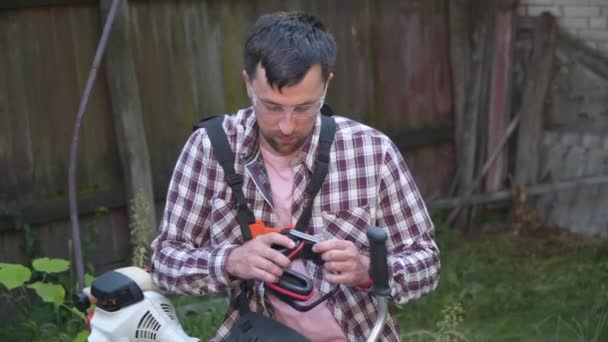 Der junge kaukasische Bauer schneidet mit einer benzinbetriebenen elektrischen Sense Unkraut, hohes Gras, Büsche und Unkraut aus dem Hinterhof des Bauernhauses. Mann arbeitet im Dorf mit Rasenmäher Gartenkonzept — Stockvideo