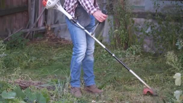 Gardener works in yard of rural house with lawn mower. Male farmer cutting wild grass with an electric scythe at the farm. Young male worker cutting grass weeds, using trimmer. Gasoline brush cutter — Stock Video