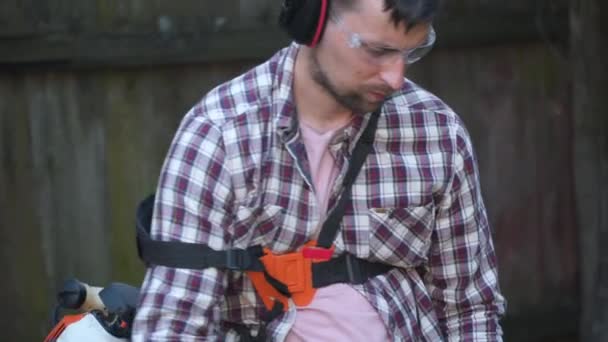 Young Caucasian farmer uses gasoline powered electric scythe to trim weeds, tall grass, bushes and weeds from farmhouse backyard. Man works with lawn mower in village. Gardening concept — Stock Video