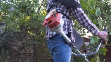 Elektrikli çim biçme makinesi ve çim biçme makinesi olan bir adam. Bahçıvan bahçe buduyor. Banliyö, köy. Beyaz tenli erkek bahçıvan avluda benzin makası ile dikiliyor. Tarlada yabani otları kesen fırça kesiciler