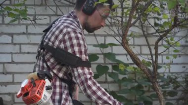 Bahçıvan kırsal evin bahçesinde çim biçme makinesiyle çalışıyor. Çiftlikte elektrikli tırpanla yabani otları kesen erkek çiftçi. Genç erkek işçi çim biçme makinesi kullanarak çim biçiyor. Benzin fırçası kesici.