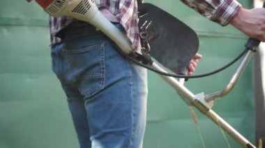 Elektrikli çim biçme makinesi ve çim biçme makinesi olan bir adam. Bahçıvan bahçe buduyor. Banliyö, köy. Beyaz tenli erkek bahçıvan avluda benzin makası ile dikiliyor. Tarlada yabani otları kesen fırça kesiciler