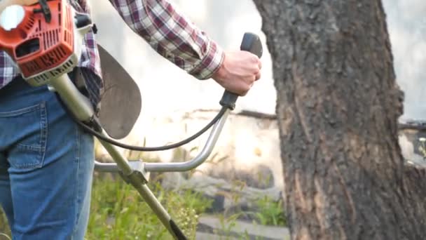 Ung kaukasisk bonde använder bensindriven elektrisk lie för att trimma ogräs, högt gräs, buskar och ogräs från gårdens bakgård. Man arbetar med gräsklippare i byn. Trädgårdskoncept — Stockvideo