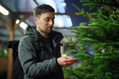 Genç Kafkasyalı adam yılbaşı tatilinde evini dekore etmek için Noel pazarından kozalaklı ağaç seçiyor. Tatil alışverişi. Erkek, akşamları dışarıdaki bir siteden Noel ağacı alır..
