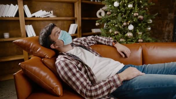 Young man in mask lies on sofa and stares at ceiling, suffering from loneliness and home isolation during Christmas holidays, coronavirus quarantine. Male suffering from boredom looking up covid 19 — Stock Video