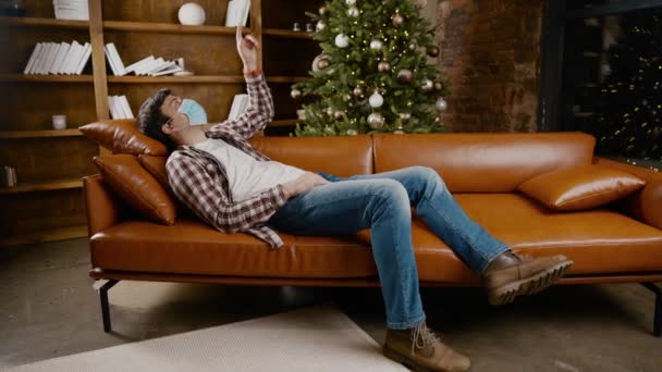 Ковид-19 и пандемия Новой Нормальной концепции. Молодой человек в маске лежит на диване один, перед рождественской ёлкой. Коронавирус накануне Рождества. Депрессивный мужчина в медицинской маске дома — стоковое видео