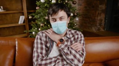 Yılbaşı için evde, kanepede maske takmış depresif bir adam var. Noel arifesinde Coronavirus. 19. mahkumiyet ve karantina döneminde arkadaşsız ve ailesiz yeni yıl tatili.