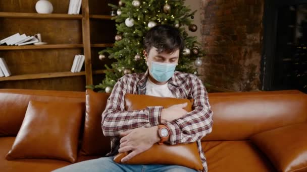 Zdenerwowany złym humorem człowiek w masce ochronnej cierpi z powodu samotności w domu podczas siedzenia na kanapie w pobliżu choinki podczas wakacji noworocznych. Nowa norma, zamknięcie i kwarantanna koronawirusów — Wideo stockowe