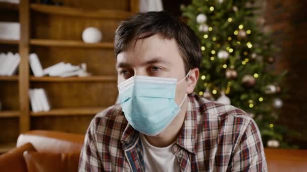 保護マスクの悪い気分の男と動揺は、クリスマスツリーの近くのソファに新年の休暇中に座っている間、自宅で孤独を患っています。新しいノルム、ロックダウンと隔離コロナウイルス — ストック動画