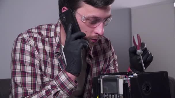 Young Caucasian man service engineer repairs kitchen equipment at customers home, clarifies availability of parts by phone, holds mobile phone and tool hands, wearing protective glasses and gloves — Stock Video