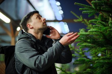 Şenlikli alışveriş, seçim ve Yeni Yıl nitelikleri satın alma. Beyaz adam telefonda konuşuyor ve yılbaşı arifesinde Noel ağacını seçmeyi tavsiye ediyor. Erkek ladin satın alır ve akıllı telefon kullanır..