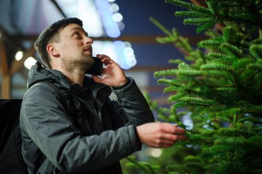 Şenlikli alışveriş, seçim ve Yeni Yıl nitelikleri satın alma. Beyaz adam telefonda konuşuyor ve yılbaşı arifesinde Noel ağacını seçmeyi tavsiye ediyor. Erkek ladin satın alır ve akıllı telefon kullanır..