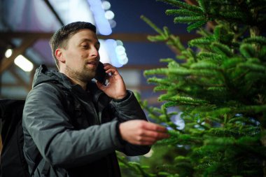 Şenlikli alışveriş, seçim ve Yeni Yıl nitelikleri satın alma. Beyaz adam telefonda konuşuyor ve yılbaşı arifesinde Noel ağacını seçmeyi tavsiye ediyor. Erkek ladin satın alır ve akıllı telefon kullanır..